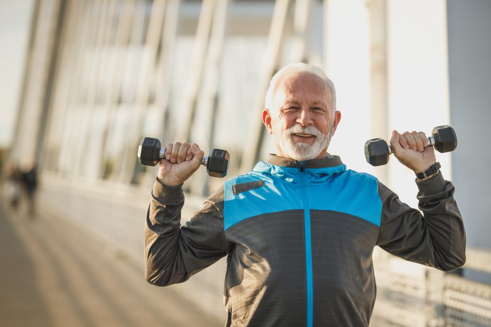 Older man smiling while lifting dumbbells overhead outdoors